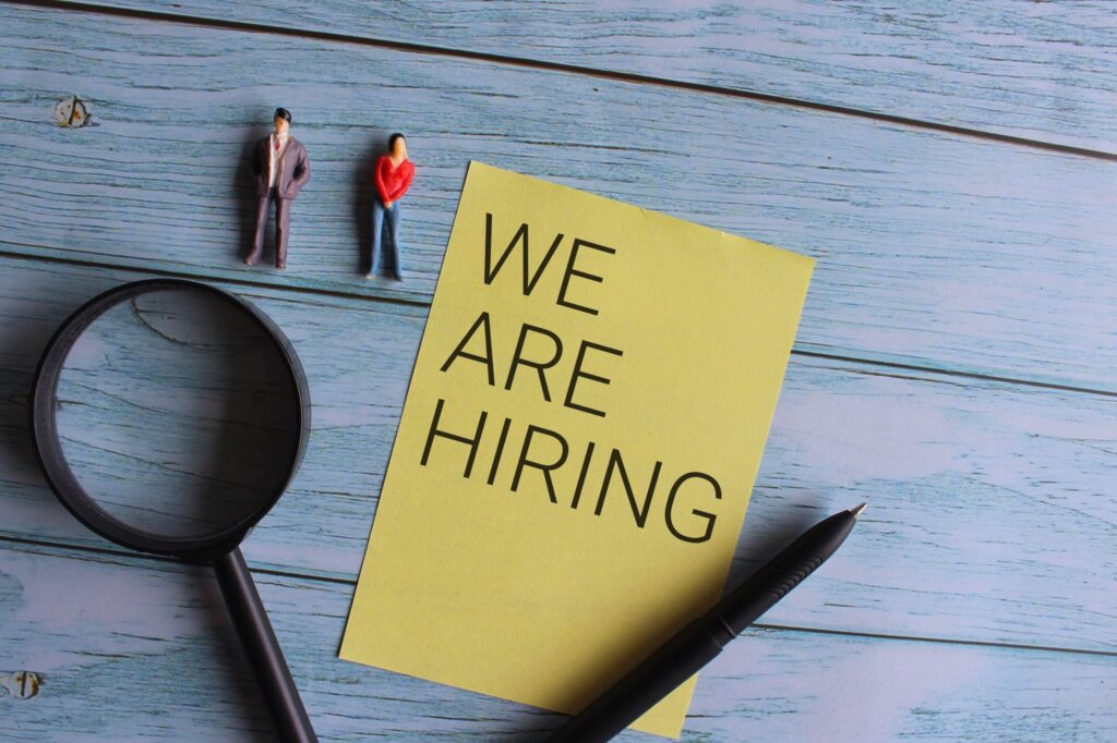 Top view of miniature people and magnifying glass with text WE ARE HIRING