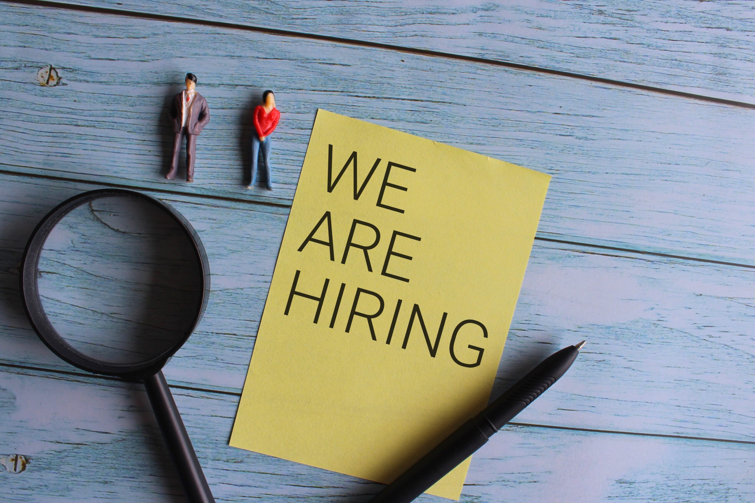 Top view of miniature people and magnifying glass with text WE ARE HIRING